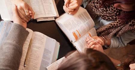 A group of young women bow their heads and pray with bibles.